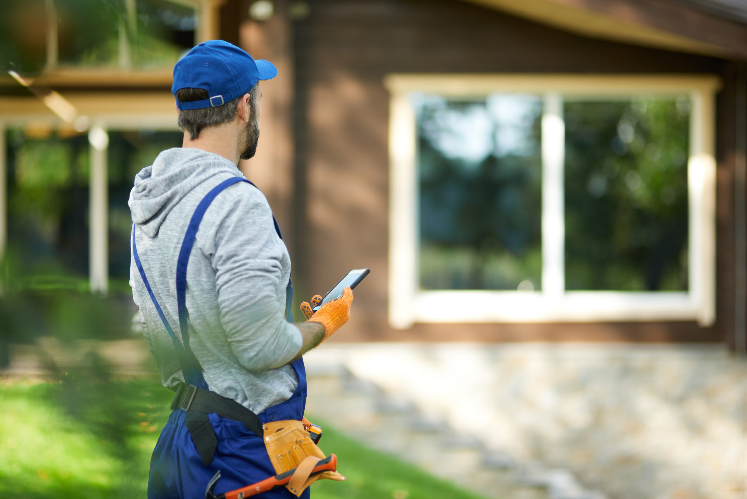 Young male builder in uniform using smartphone, standing outdoors while working at cottage construction site on a sunny day